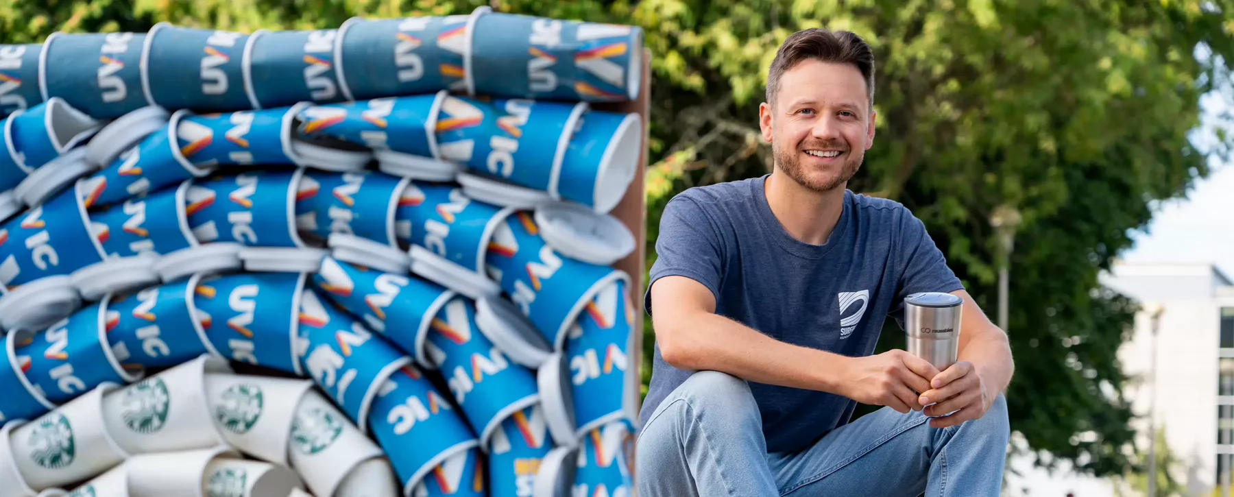 Student sits behind a colourful collage of UVic single-use beverage cups. He is holding a metal reusable coffee tumbler and wearing a navy blue t-shirt.