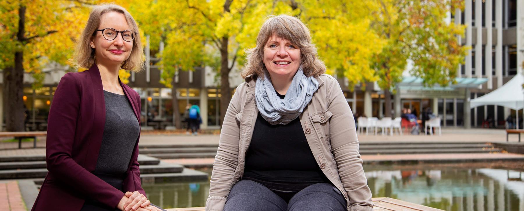 Professors Erin E. Kelly and Sara Humphreys sit next to the fountain outside of UVic Library