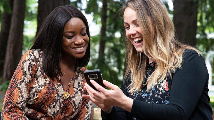 Two women sit on a bench laughing while looking at a cell phone.