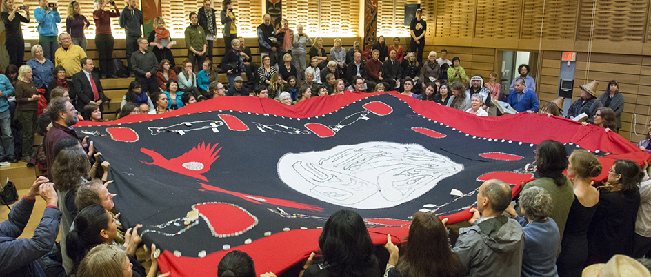 A group of people in the First Peoples' House holding a button blanket
