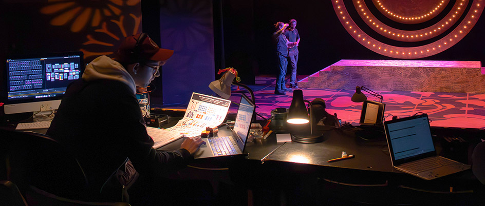 Students working on a computer while others set up lights on a stage