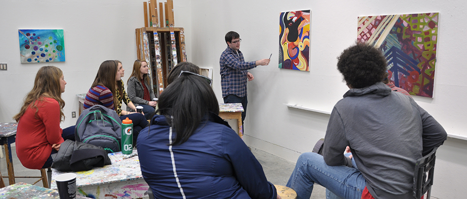 Students listening to an instructor in an art classroom while he gestures at a painting