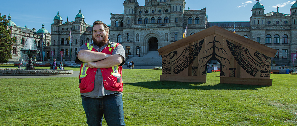 Tlehpik Hjalmer Wenstob standing with his Indigenous art installation on the law of the BC Legislature Buildings