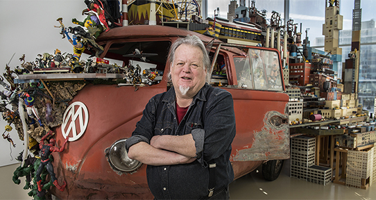 Artist and alumnus Kim Adams standing in front of his sculpture featuring a Volkswagen.