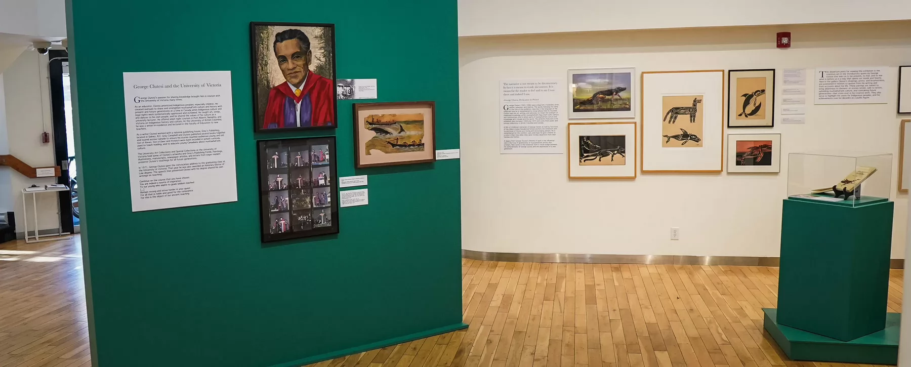 A display in an art gallery containing a painting of an Indigenous man in a graduation robe, framed art work and a text card titled George Clutesi and the University of Victoria.