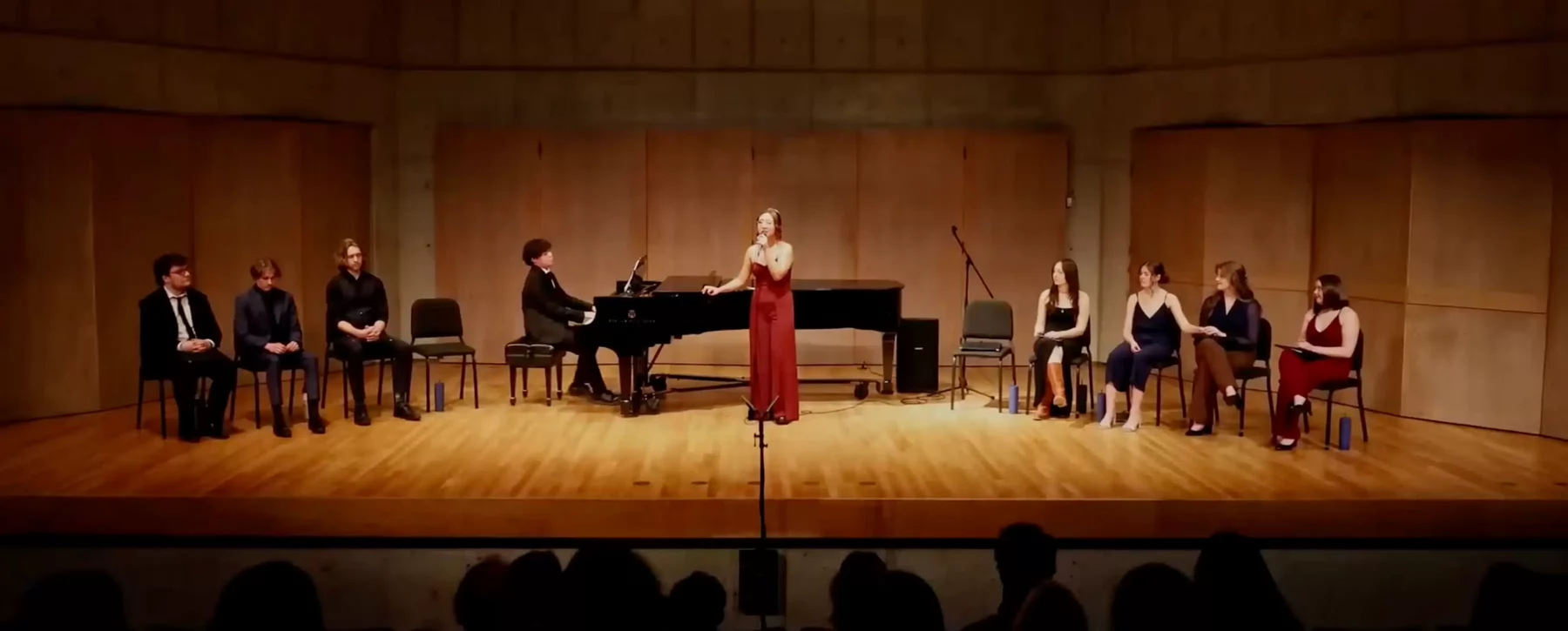 A singer standing on stage performing at a recital beside a piano and students seated on stage. 