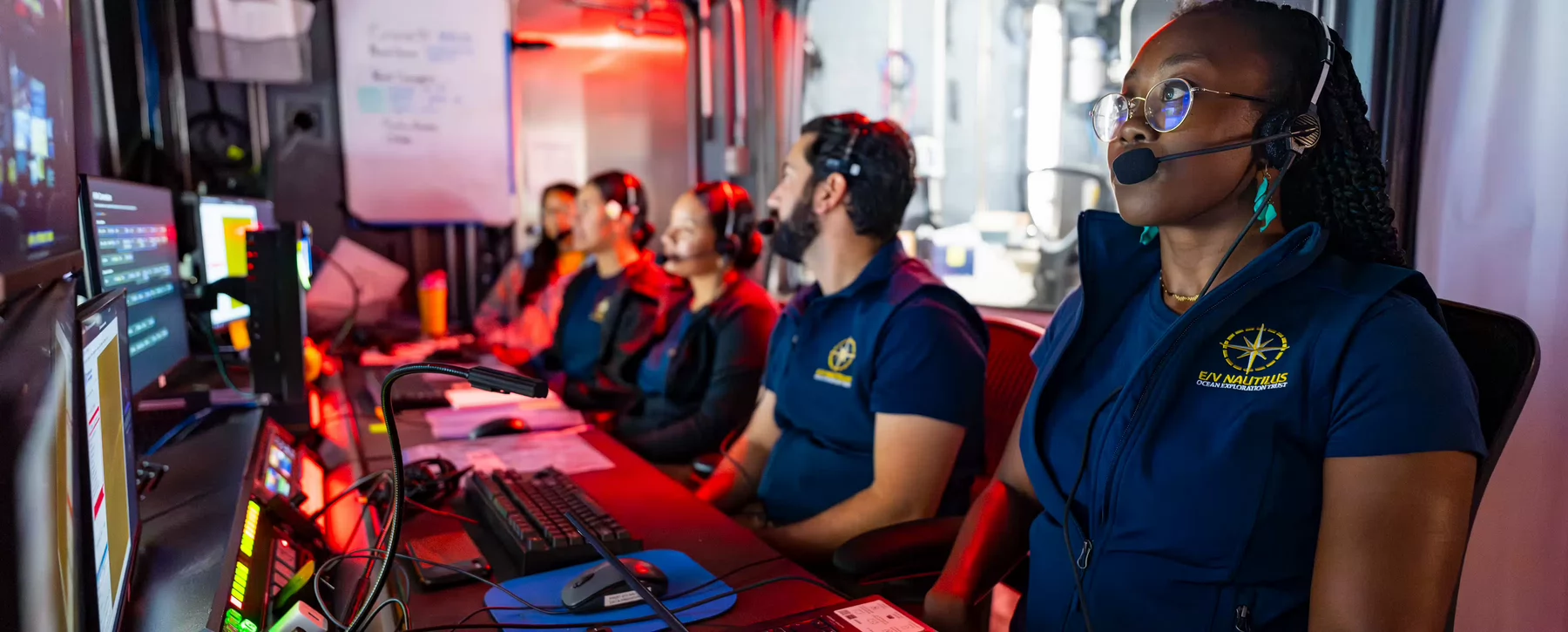 Woman with braided hair, glasses and headset microphone looks at a computer screen at a table with four other people with headset microphones looking at computer screens. 