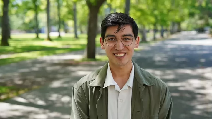 A male student with dark hair and glasses standing on a tree covered university campus.