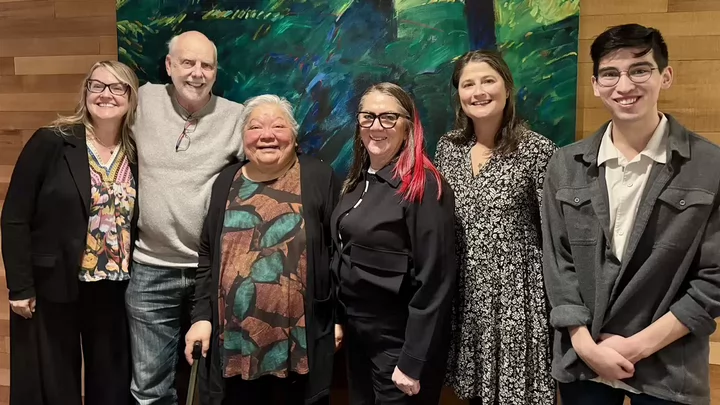 Six people standing shoulder to shoulder smiling in front of a colourful backdrop.