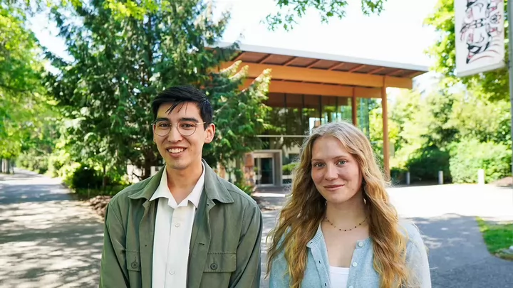 A male student with dark hair and glasses standing beside a female student with long blond hair outside in front of a building surrounded by trees.. 