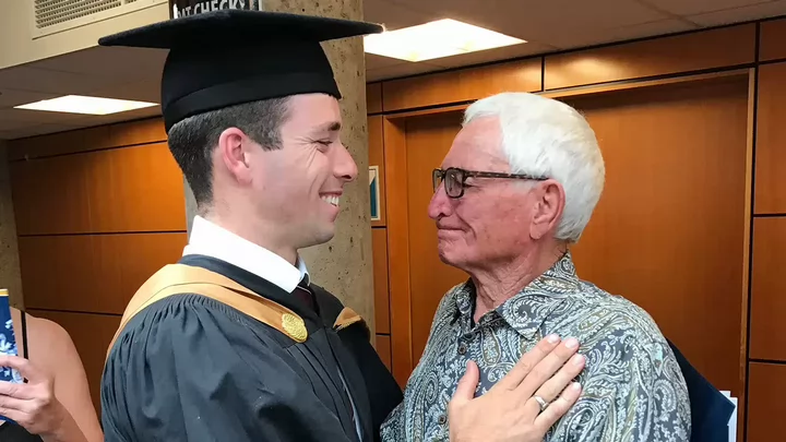 A young man wearing a graduation robe and cap smiling at an older man.