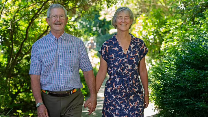 Man with glasses, moustache and buttoned up shirts holding hands with woman wearing a flower patterned dress and smiling.