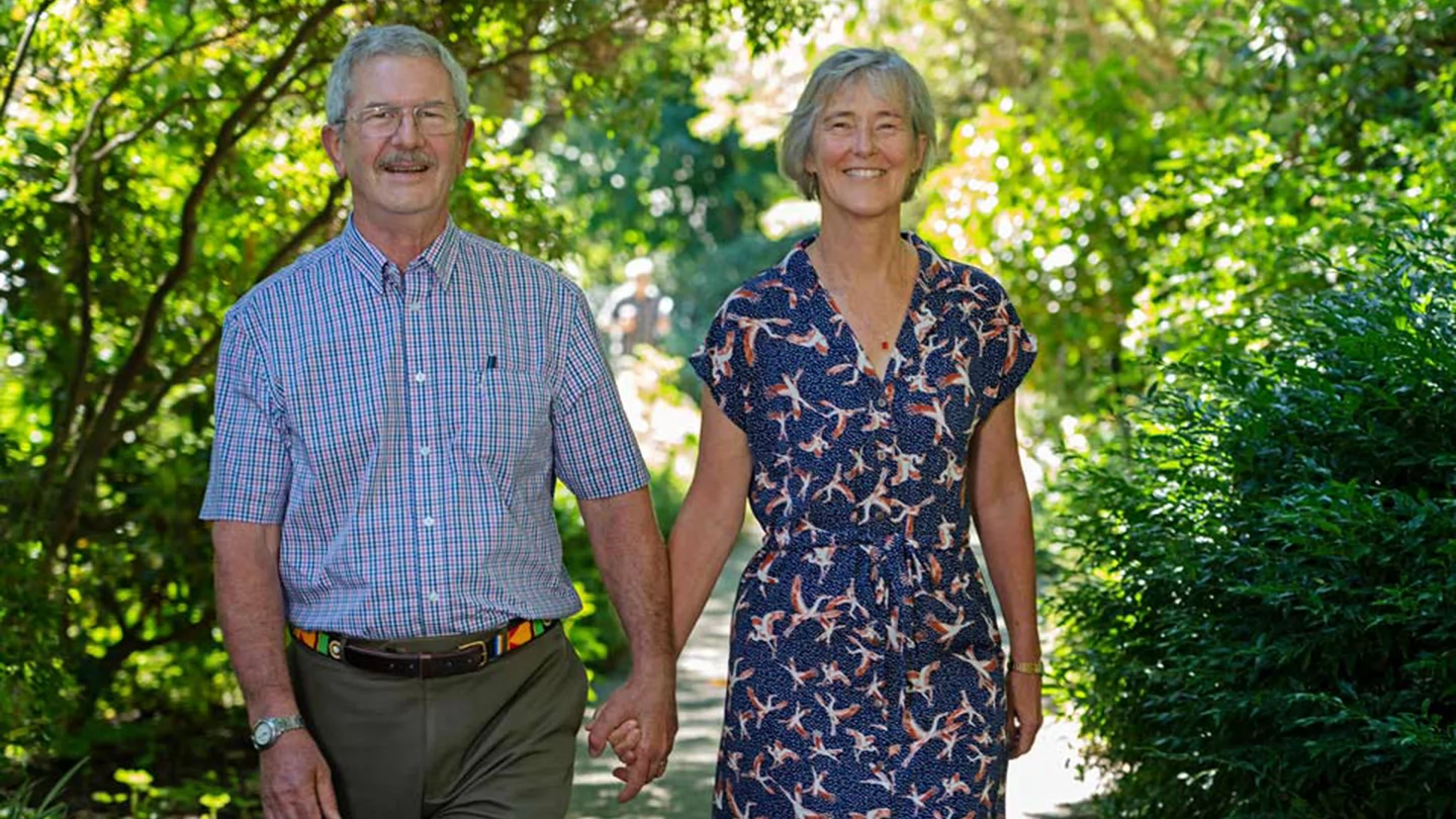 Man with glasses, moustache and buttoned up shirts holding hands with woman wearing a flower patterned dress and smiling. 