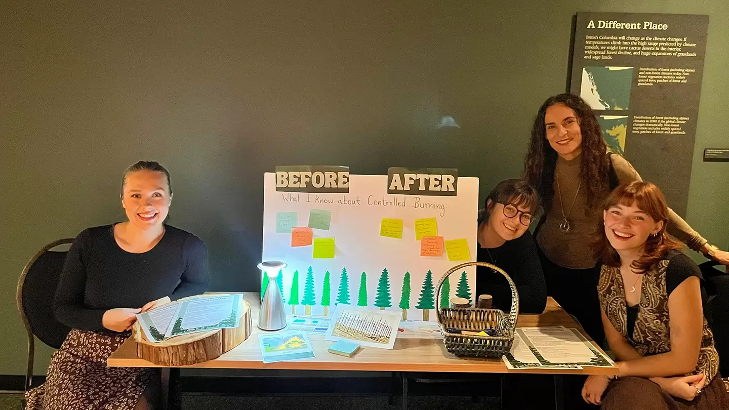 Erika Germanos stands with three undergraduate students at a learning station about controlled burning at the Royal BC Museum.