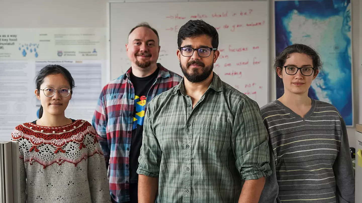 Four people standing shoulder to shoulder in an office with a map and a white board filled with questions in the background. 