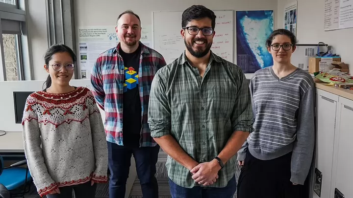 Four people standing side by side in an office space with white boards and a map in the background. 