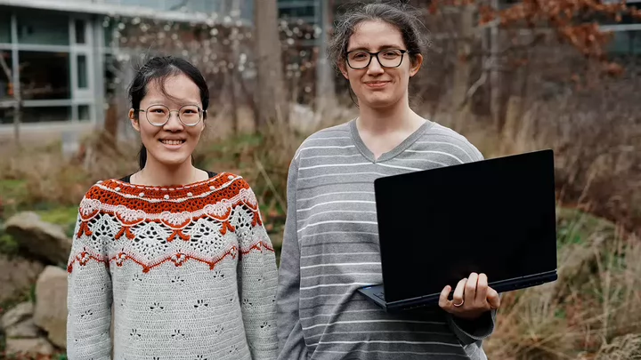 Two students wearing glasses standing side by side outside and one is holding a laptop computer.
