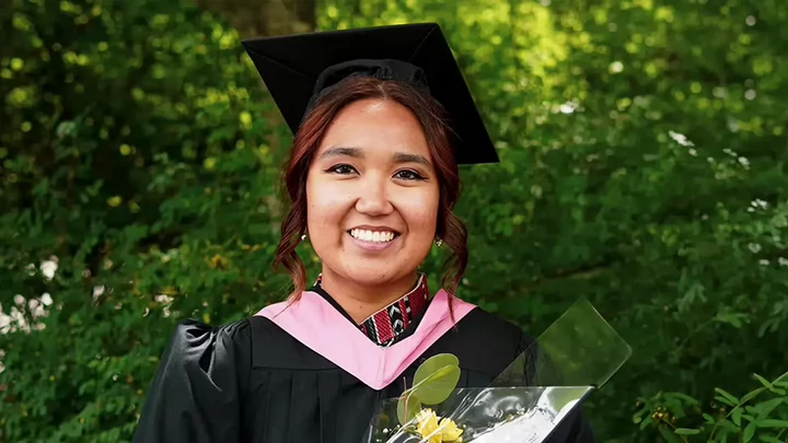 A university graduate in cap and gown holding flowers and smiling. 