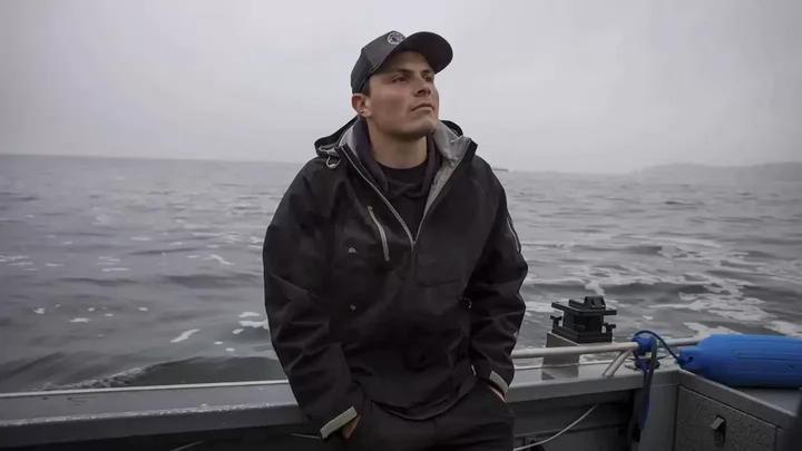 Man wearing a windbreaker jacket and baseball hat standing on a boat floating on the ocean.
