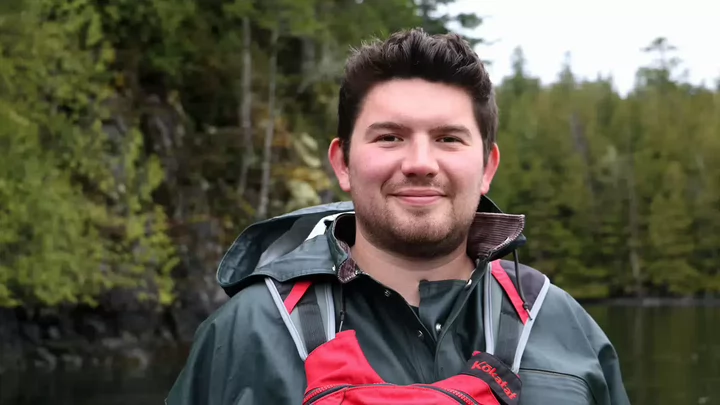 A man with facial hair and a raincoat standing in a forested area surrounding a river..