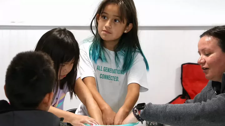 Three children, one wearing a T-shirt that reads All Generations Connected, and a teacher placing their hands in a bowl.