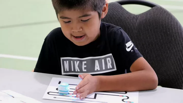 A child is seated at a table and guides an “Ozobot” along a path drawn on a piece of paper.