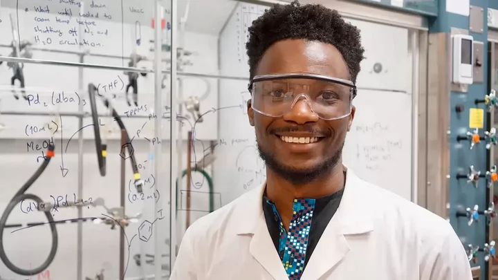 Man with facial hair, wearing safety goggles and a lab coat smiling in front of a white board covered with scientific formulas.