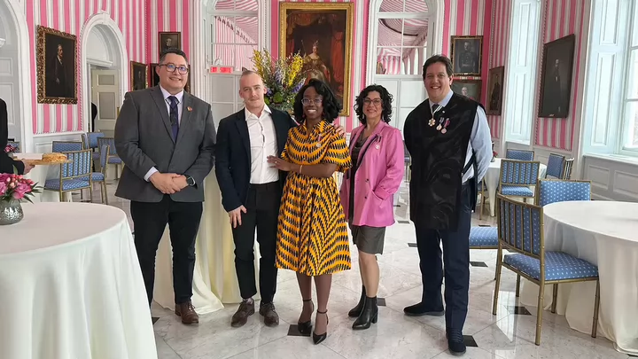 Four people smiling and posing for a photo in an ornately decorated room will with painted portraits of people from long ago.