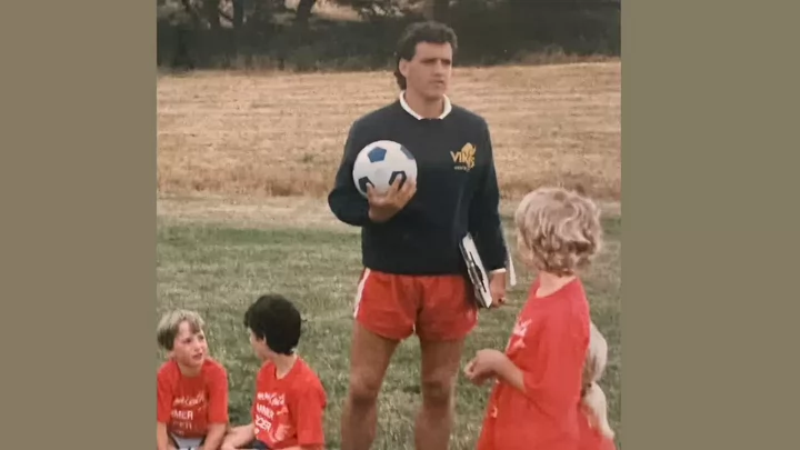 A young man wearing shorts and a sweatshirt that says Vikes holding a soccer ball while instructing kids at a soccer camp.