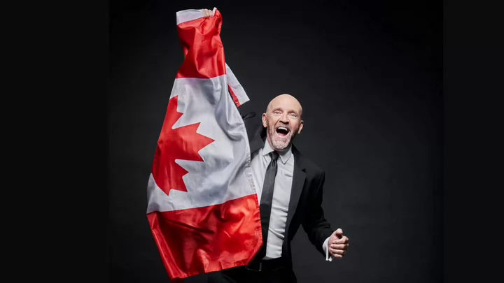 A man wearing a suit and tie cheering while holding up a large Canadian flag.