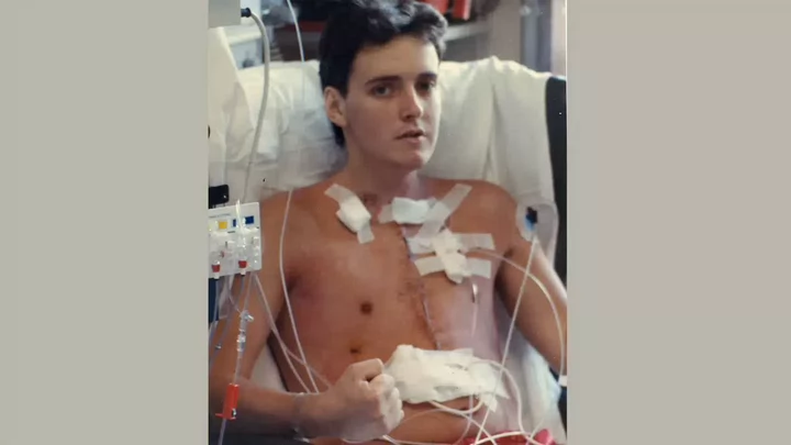 An older photo of a young man in a hospital bed hooked up to wires and tubes and stitches down the middle of his chest.