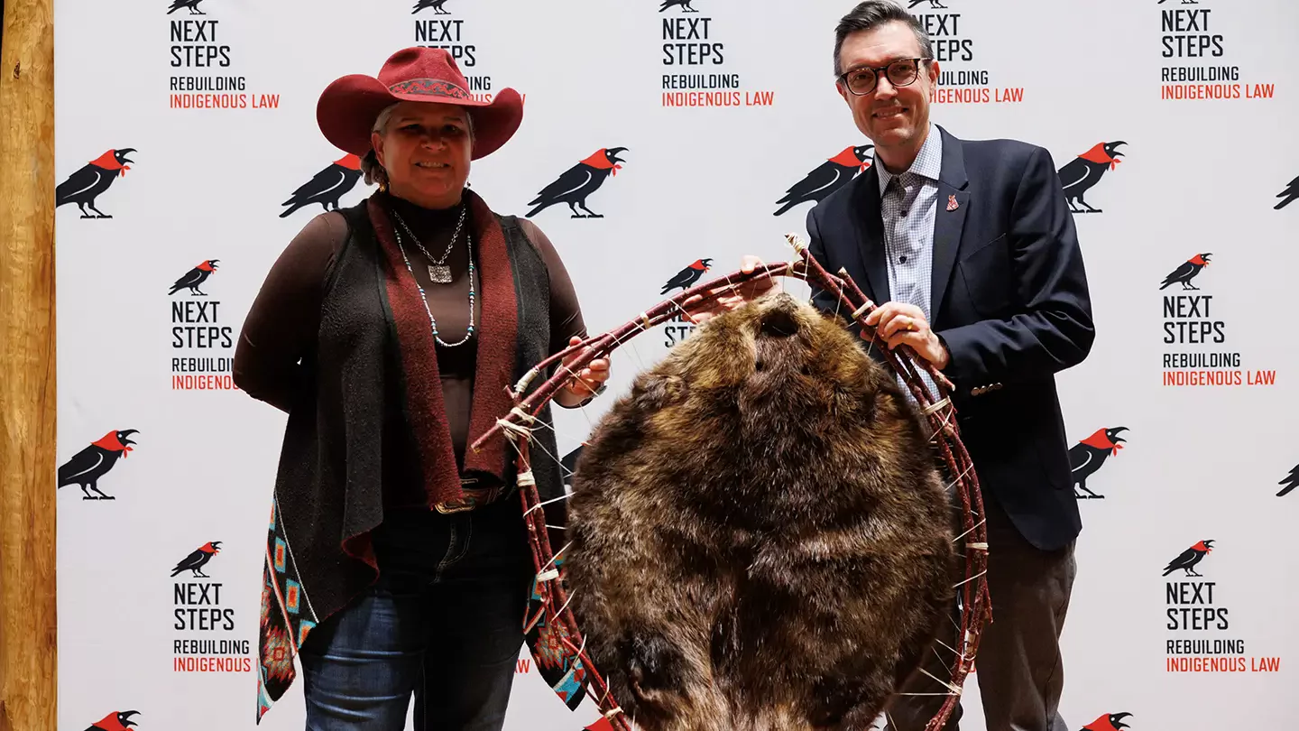 A person wearing a red hat and a person in a suit hold a beaver pelt hung on a large willow hoop between them