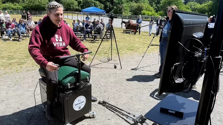 A person sitting astride the Miracolt horse riding simulator opposite a tv screen, with a crowd of people in the background