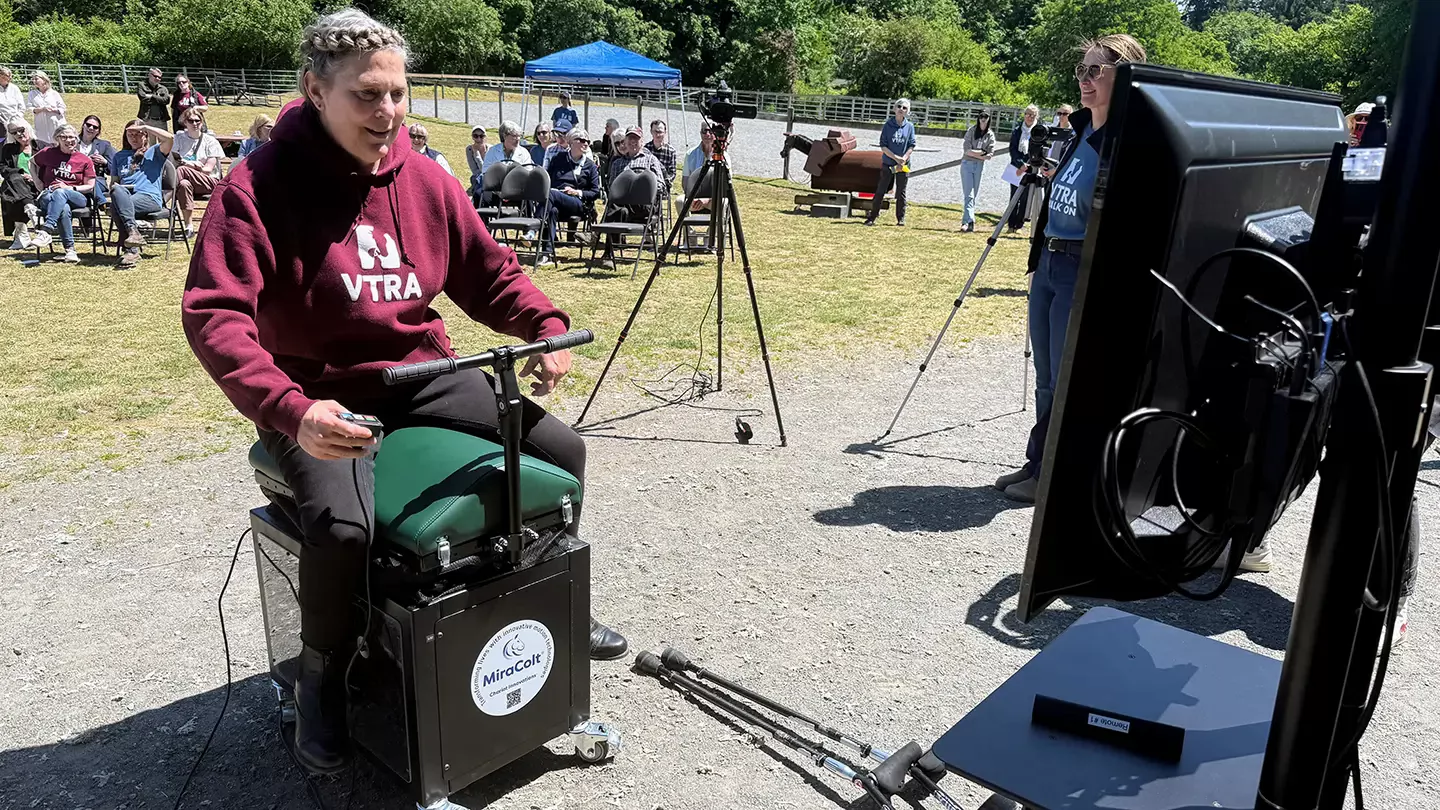 A person sitting astride the Miracolt horse riding simulator opposite a tv screen, with a crowd of people in the background