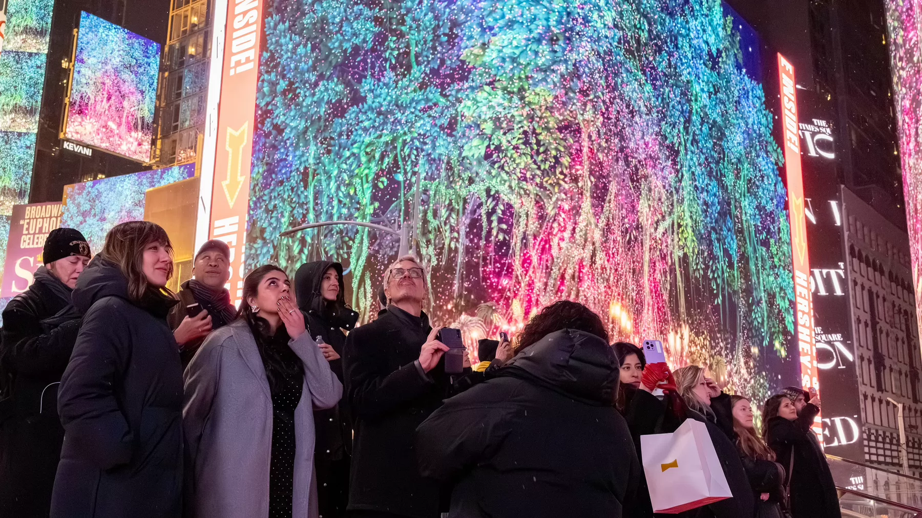 A group of people standing on a street corner looking up at a digital image of a tree on a digital billboard in New York City's Times Square.