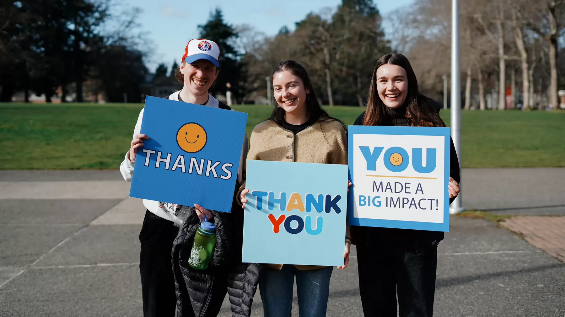 Three students standing side by side smiling and holding signs that read "Thanks," "Thank You" and "You Made a Big Impact."