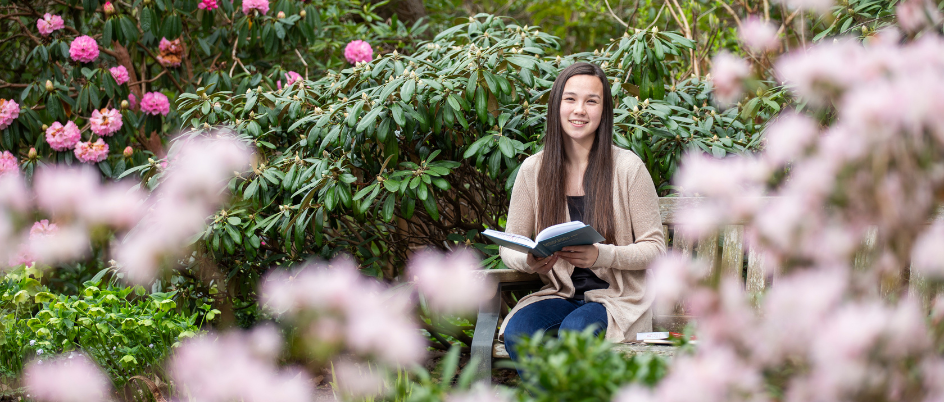 Arista Marthyman, a young female student, smiles at the camera while sitting in Finnerty Gardens, she is sitting on a bench holding a blue book open and pink flowers are in view. 