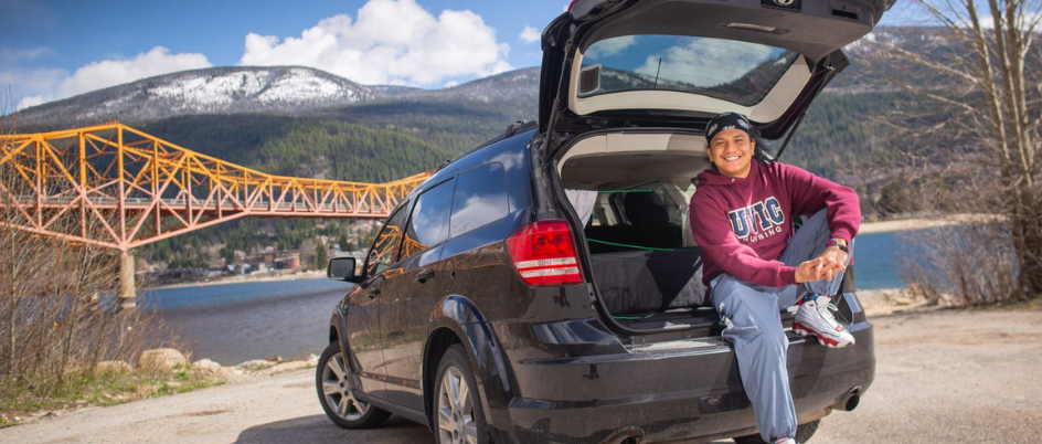James Matthew Besa smiles while sitting in the trunk of a black car’s trunk.  James is wearing a red UVic sweater. There is a river and mountains in the background.