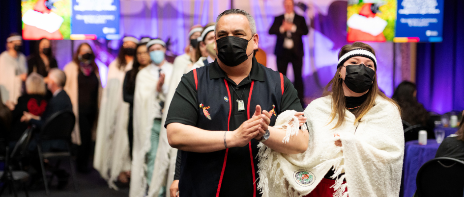 An Indigenous man, Sheshatshiu Innu First Nation Chief Eugene (left) and Innu woman, Jolene Ashini (right), walk arm in arm smiling beneath black masks. Jolene is wearing Indigenous regalia with a white braided headband with black trim, a cream blanket with Indigenous artwork on the emblem and red skirt. 