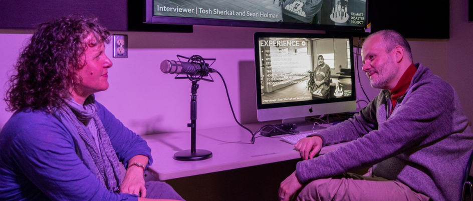 Sean Holman (right), sits in a studio facing Sandra Ibrahim (left). Both are looking at each other and smiling with screens on behind them that read “climate disaster project” on one screen and a photo of a woman in black and white in an interview on the other. The room has a purple hue to it. 