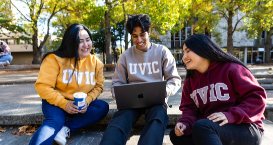 three students wearing uvic sweaters sit on steps and smile looking at a laptop in the middle