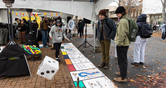 Three students are in focus playing a life-sized monopoly style game with a massive dice in mid-air