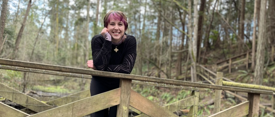 Student with short cropped pink and brown hair, wearing all black smiling at the camera while standing on a bridge in the forest.
