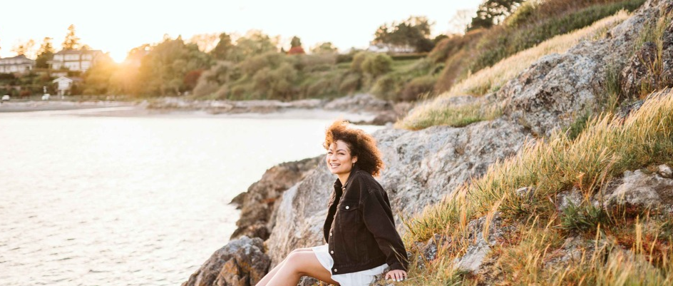 Meghan Wilson sits on rocks in front of the ocean smiling at the camera