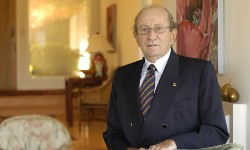 Man in suit standing with hands resting on a chair back in a living room