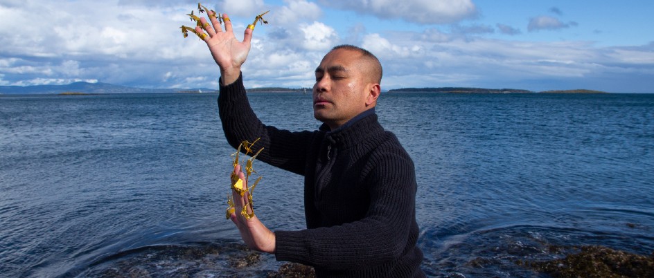 Male dancer wearing golden finger adornments posing on a rock by the ocean.