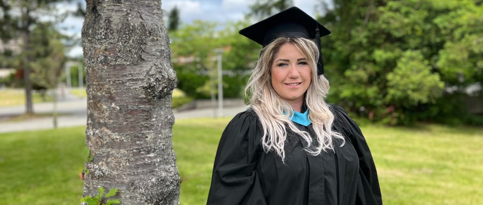 Student in convocation attire stands by a tree on UVic campus.