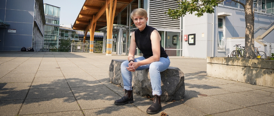 Student with short cropped blond hair, wearing black tank, jeans and boots sits on stone bench in front of campus building. 