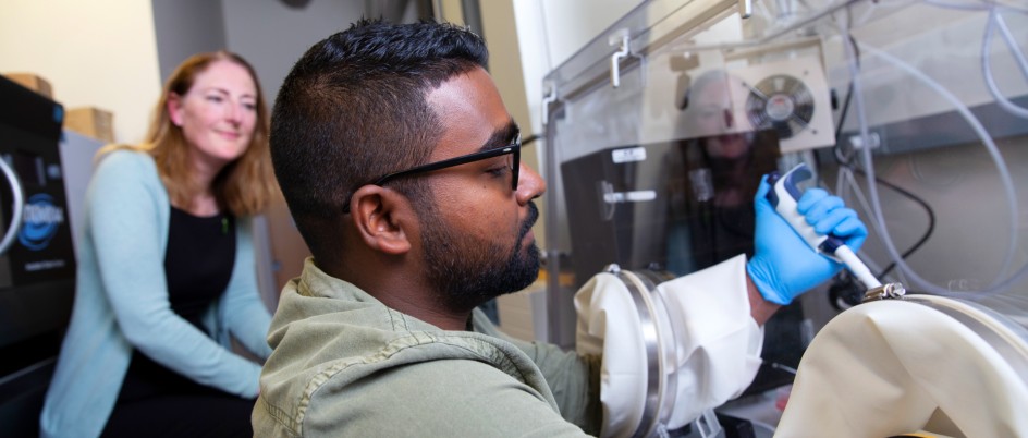 Student researcher leans over a microscope as supervisor looks over his shoulder