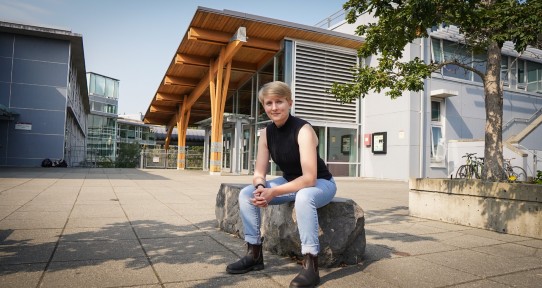 Student with cropped blond hair and blue eyes sitting on stone bench in front of university building.